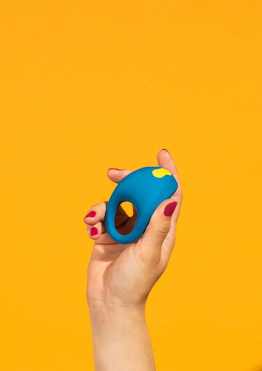 Hand with red nail polish holding a blue silicone cock ring against a bright yellow background