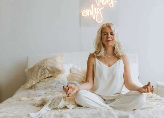 Older woman with gray hair meditating cross-legged on bed in bright, cozy bedroom