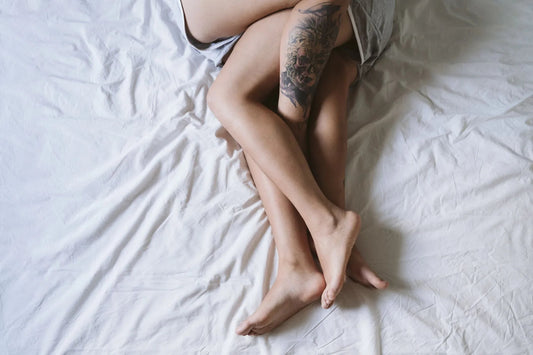 Two pairs of intertwined bare legs on white bed sheets, one leg with a detailed tattoo